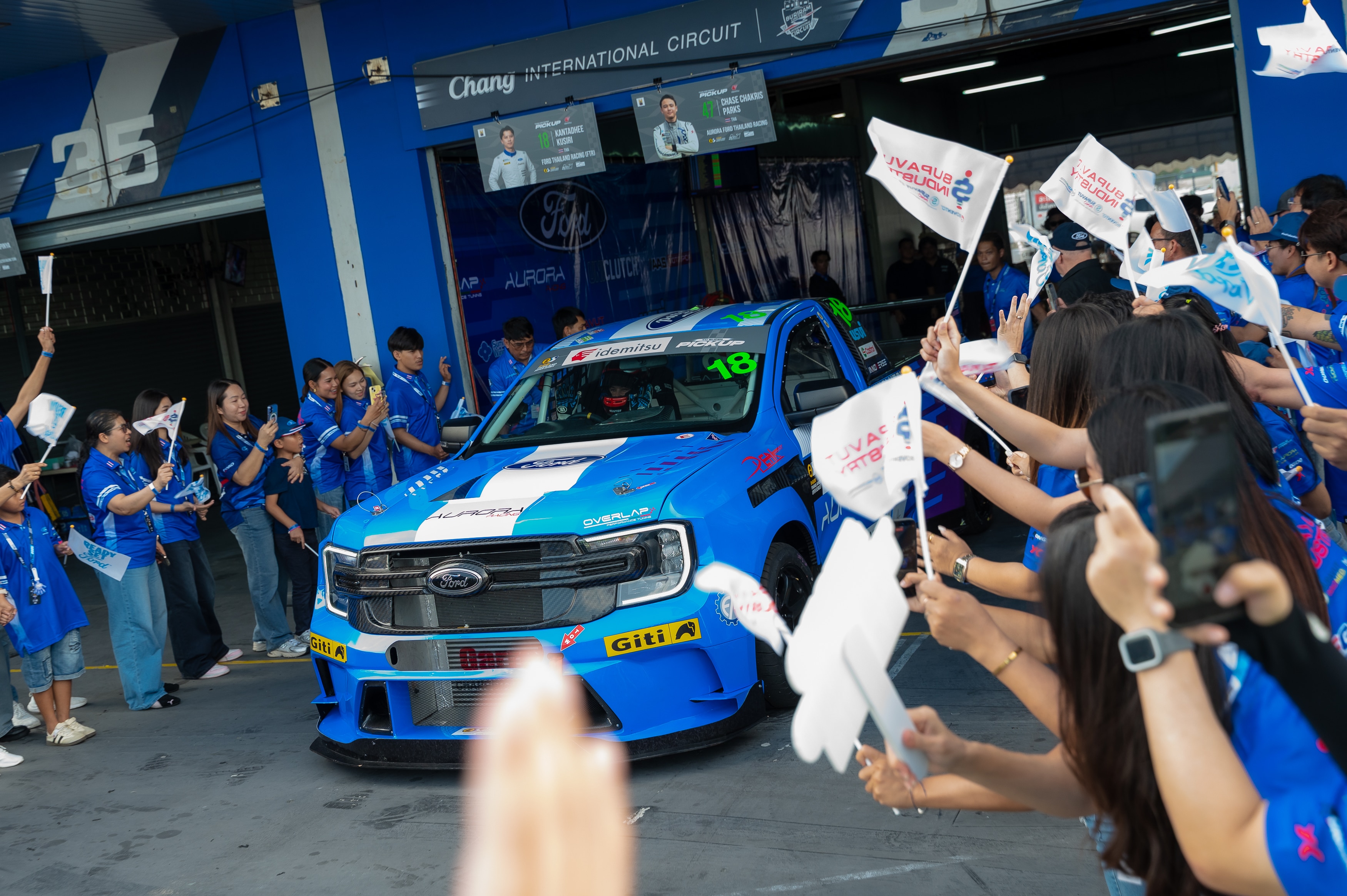 A crowd of excited fans in blue Ford Thailand Racing apparel cheering as a Ford Ranger race truck exits its garage bay.